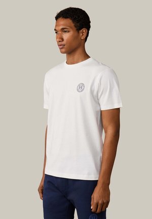 Homme portant un t-shirt blanc à manches courtes avec un petit logo circulaire sur la poitrine et un pantalon bleu marine, se tenant devant un fond clair uni.