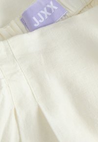 Vêtement de couleur crème avec des coutures visibles et une étiquette en tissu lavande pliée affichant le nom de la marque "JUXX".