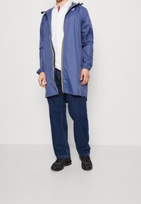 Lang blå regnjakke med hætte, lynlås, elastiske manchetter og gul lynlåsdetalje, båret over en hvid skjorte med krave og blå jeans.