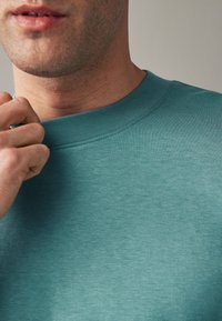 Türkiser Rundhals-Sweatshirt mit weicher Textur und geripptem Kragen. Nahansicht von Stoff- und Nähdetails.