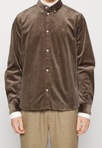 Camisa de pana marrón con cuello abotonado, botones blancos y sutiles detalles bordados. Mangas largas con puños en contraste.