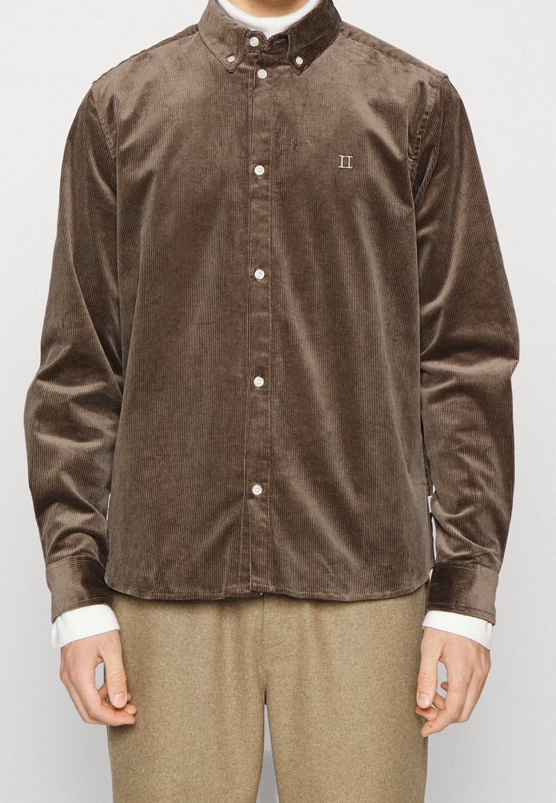 Camisa de pana marrón con cuello abotonado, botones blancos y sutiles detalles bordados. Mangas largas con puños en contraste.