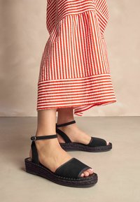 Zwarte suède wedge sandalen met een open teen, enkelbandje en een gestructureerd espadrille platform. Gecombineerd met een rood-wit gestreepte rok.