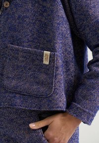 Blaue Wollmischungjacke mit Fischgrätenmuster, mit braunen Akzenten, einer Brusttasche und einem Etikett am Tascheneingang.