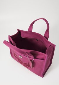 Różowa kanwasowa torba na ramię z podwójnymi uchwytami, wewnętrzną kieszenią na zamek, wytłoczonym logo z przodu oraz oddzielną zawieszką na łańcuszku.