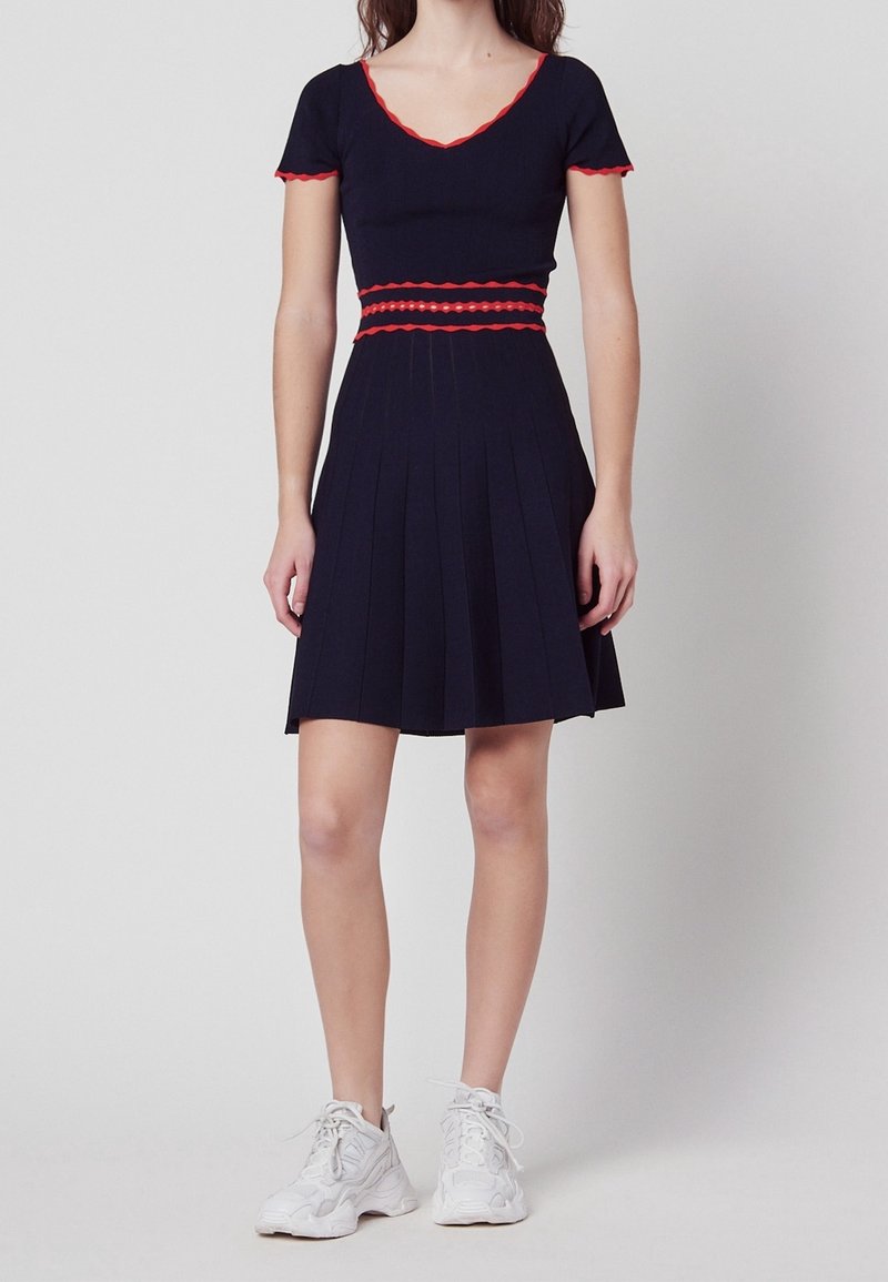 Marindräkt med korta ärmar, röd vågig kant, figurnära liv och plisserad kjol. Bärs med vita sportskor.