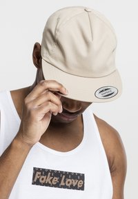 Beige snapback pet van stof, met een gebogen klep en een logo-sticker aan de voorkant. Draagt door een persoon in een witte tanktop.
