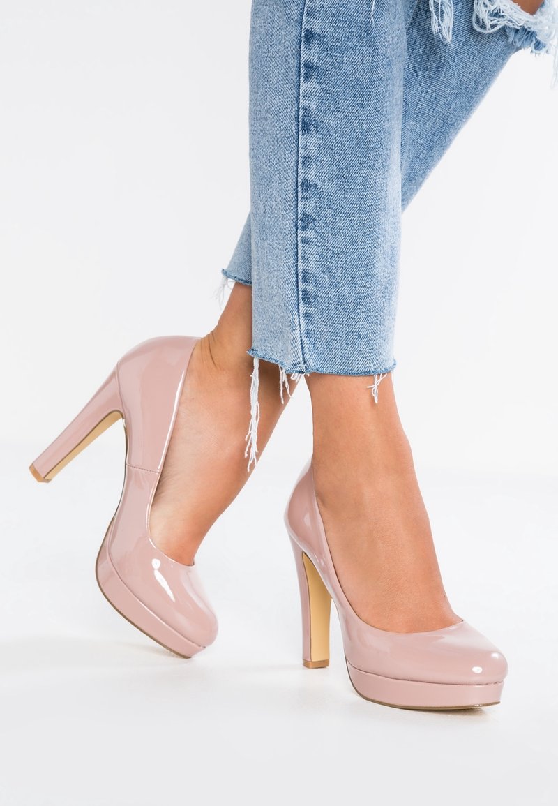 Jambes en jean déchiré bleu clair portant des chaussures à plateforme à talons hauts en cuir verni nude brillant sur fond blanc.