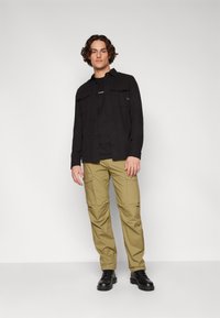 Μαύρο πουκάμισο με μακριά μανίκια και κλείσιμο με κουμπιά, χακί cargo παντελόνι με πλαινές τσέπες και μαύρες μπότες με κορδόνια. Απλός και λειτουργικός σχεδιασμός.