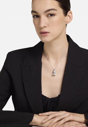 Collar plateado con un colgante en forma de oso, llevado sobre una blusa negra con escote en V. El blazer es negro con hombros estructurados.