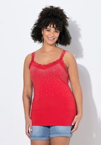 Top rosso senza maniche con rifiniture in pizzo, decorato con borchie argentate che sfumano dal alto verso il basso. Abbinato a pantaloni corti in denim arrotolati.