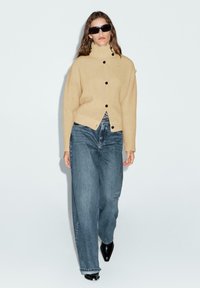 Beige ribbad tröja med hög krage och svarta knappar, ihop med lösa blå jeans och svarta spetsiga skor. Modellen går framåt.