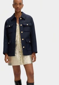 Mörkblå jacka i ullblandning med guldknappar och fyra frispårsfickor, bärs över en glänsande guldfärgad metallisk miniklänning.