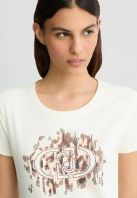 Crème kleurige t-shirt met een ronde halslijn, voorzien van een centraal ontwerp van kleine, metalen studs in brons en zilver die een logopatroon vormen.