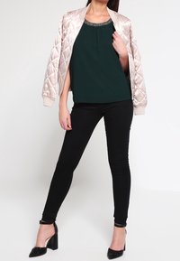 Donna con una giacca bomber beige trapuntata lucida sopra un top verde scuro, pantaloni skinny neri e scarpe con tacco a blocco nere, in piedi con una gamba avanti.