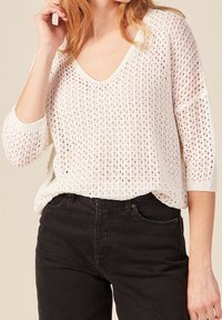Bílý pletený svetr s volným střihem, s výstřihem ve tvaru V a tříčtvrtečními rukávy. Nosený s černými džíny s vysokým pasem.