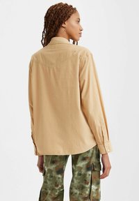 Camicia a maniche lunghe marrone chiaro con chiusura a bottone e yoke sul petto; abbinata a pantaloni cargo mimetici verdi con tasche laterali.