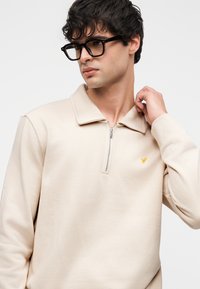 Junger Mann mit dunklen Haaren und Brille, der einen beigefarbenen Pullover mit Reißverschluss trägt, auf der Brust ein kleines gelbes gesticktes Logo, vor weißem Hintergrund.