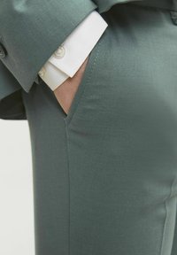Pantaloni su misura in tessuto teal. Le caratteristiche includono una superficie liscia, tasca laterale e un polsino di camicia bianco che spunta dalla manica della giacca.