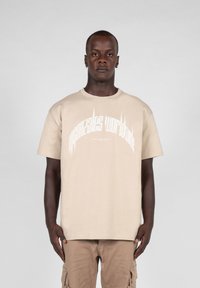 Beige bomull t-skjorte med korte ermer og rund hals, med hvit grafisk tekst sentrert på forsiden. Glatt tekstur.