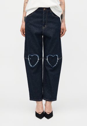 Tmavé denimové džínsy so širokými nohami a ošúchanými záplatami v tvare srdca na kolenách, nosené s čiernymi špicatými topánkami a bielym topom.