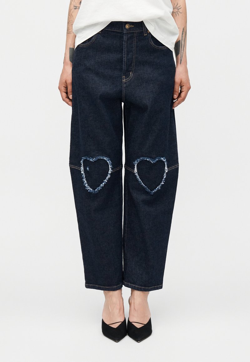 Σκούρο τζιν με φαρδιά μπατζάκια και ξεφτισμένες μπαλώματα σε σχήμα καρδιάς στα γόνατα, φορεμένα με μαύρα μυτερά παπούτσια και λευκό τοπ.