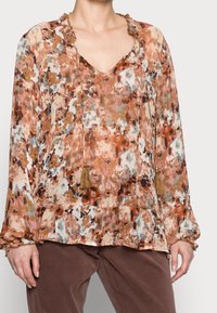 Blusa floral en tonos de marrón, naranja y crema. Presenta un escote en V, un cuello con volantes y detalles de borlas en los lazos. Tejido texturizado.