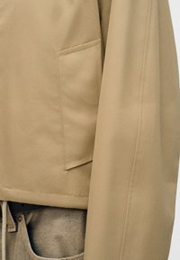 Manche et détail de poche d'une veste beige sur un jean en denim clair avec ceinture et taille visibles, mettant en valeur la texture du tissu et les coutures.