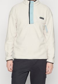 Pullover in pile color crema con colletto alto, dotato di una chiusura a snap blu, dettagli neri e una tasca sul petto con zip.