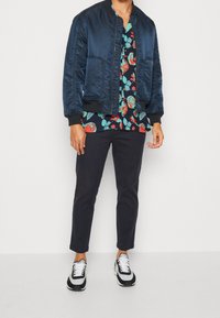 Laivastonsininen bomber-takki kukkaisella lyhythihaisella paidalla, jossa on punaisia, vihreitä ja sinisiä kuvioita; yhdistetty tummiin housuihin ja mustavalkoisiin tennareihin.