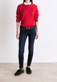 Suéter rojo de punto con un patrón texturizado, camisa blanca con cuello debajo, combinado con jeans ajustados de color azul oscuro y zapatillas negras.