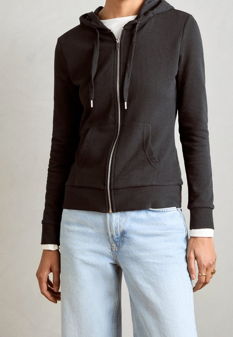 Sudadera negra con cremallera, hecha de tela suave, con capucha ajustable con cordón, bolsillos frontales y puños acanalados, combinada con jeans azul claro.