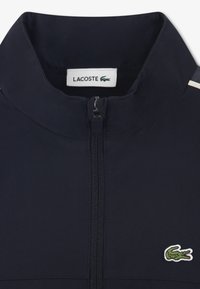 Μπλε σκούρο μπουφάν με φερμουάρ, με ε πένδυση από δίχτυ, γιακά τύπου mock και λογότυπο Lacoste στην αριστερή πλευρά. Λευκές λεπτομέρειες στους ώμους.
