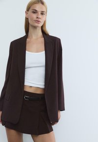 Blazer marron foncé à rayures, avec une coupe décontractée, associé à un débardeur blanc et une jupe assortie avec une ceinture noire en accent.