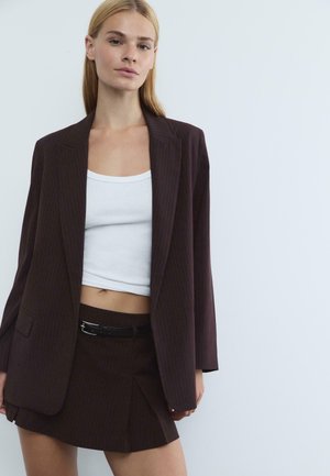 Blazer a righe pinstripe marrone scuro con una vestibilità rilassata, abbinato a una canotta bianca e a una gonna coordinata con un accento di cintura nera.