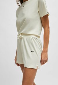 Ljusbeige kortärmad topp och shorts-set i mjukt tyg, med en svart "BOSS"-logotyp på båda delarna. Shortsen har sidoklipp.