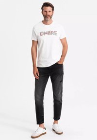 Camiseta blanca con un diseño gráfico, combinada con jeans negros desgastados y zapatillas blancas. El conjunto es casual y minimalista.
