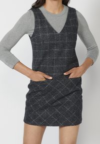 V-neck sleeveless dress in navy tweed, featuring a subtle check pattern, with a gray long-sleeve top underneath and side pockets.