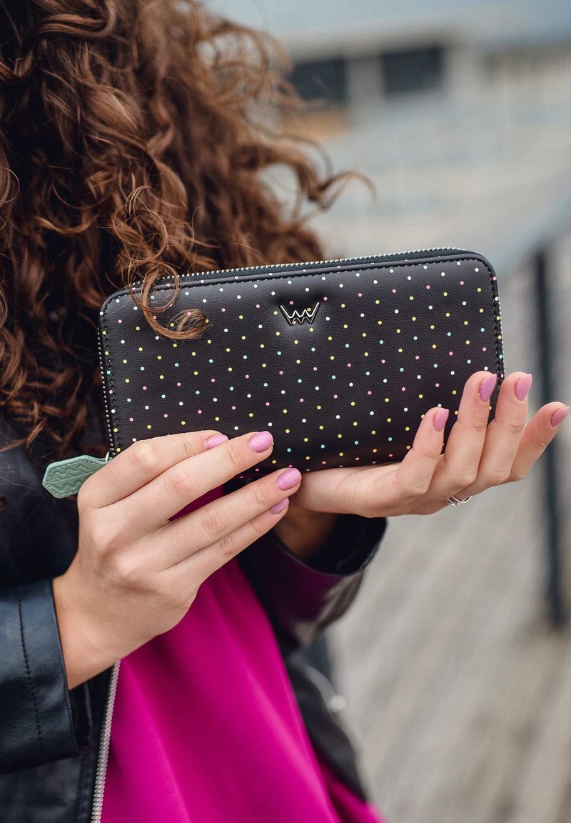 Black wallet with multicolored dots, zip closure, and green accent tag. Presented in hands with light pink nails, textured finish.