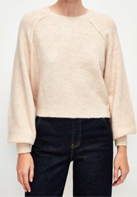 Femme portant un pull beige clair en matière pelucheuse avec des manches bouffantes, associé à un jean taille haute bleu foncé, sur un fond uni.