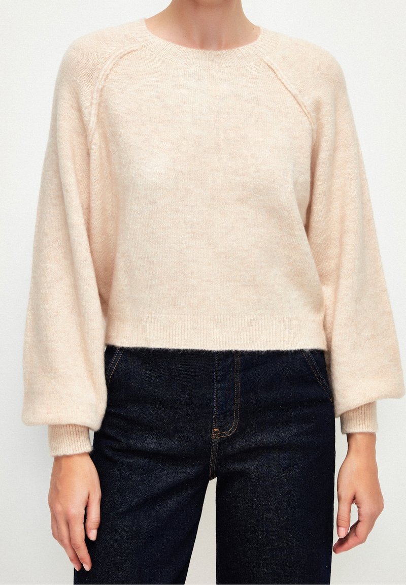 Femme portant un pull beige clair en matière pelucheuse avec des manches bouffantes, associé à un jean taille haute bleu foncé, sur un fond uni.