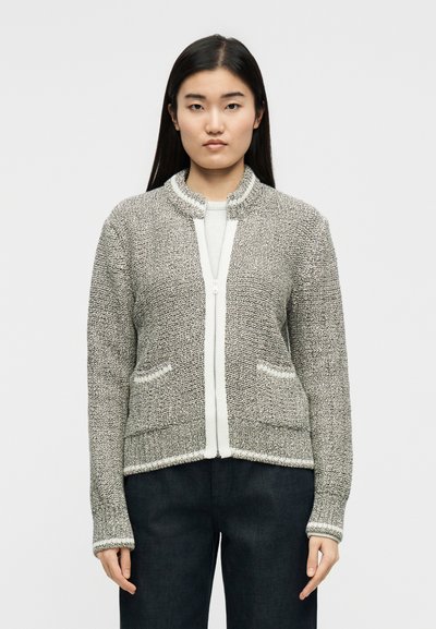 Νεαρή γυναίκα με ίσια σκούρα μαλλιά φοράει γκρι πλεκτή ζακέτα με φερμουάρ πάνω από λευκό τοπ και μαύρο παντελόνι, στέκεται μπροστά σε απλό φόντο.