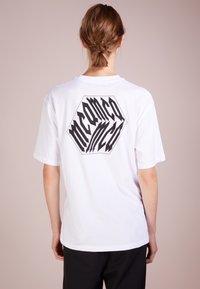 Bílé bavlněné tričko s krátkým rukávem, na zadní straně s výrazným černým hexagonálním grafickým prvkem a stylizovaným designem textu.