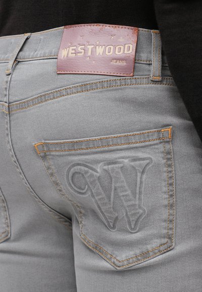 Sive traperice imaju kožni oznaku na pojasu s "WESTWOOD JEANS." Stražnji džep ima ušivan "W" logo i narančaste šavove.