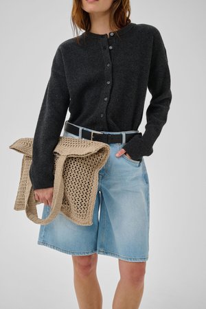 Naine, kes kannab tumedat halli nööbitavat kardigani ja hele sinist teksashäkkleid, hoides ühes käes beeži heegeldatud käekotti.