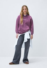 Hoodie violet avec poche avant et texte, assorti à un jean évasé gris et une chemise nouée blanche. Les chaussures sont des mocassins marron.