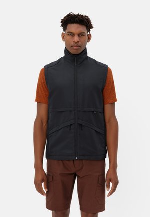 Zwarte mouwloze vest met hoge kraag, ritssluiting en voorzakken; gedragen over een oranje T-shirt met korte mouwen en een bruine korte broek.