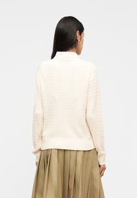 Γυναίκα με μακριά σκούρα μαλλιά, που φοράει κρεμ πουλόβερ πλεκτό με πλεξίδες και πλισέ φούστα σε ελαιόχρωμο, εμφανιζόμενη από πίσω.