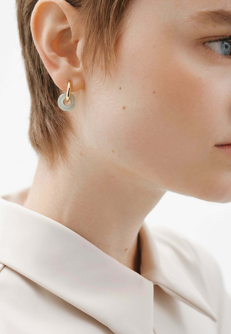 Pendientes de aro dorados con un acento circular verde translúcido. Mostrados en un modelo con cabello corto, que lleva una camisa de color claro con cuello.