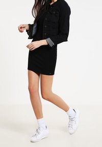 Svart denimjacka med grå foder, bärs över en åtsittande svart klänning. Vita sneakers och synliga vita strumpor fulländar outfiten.